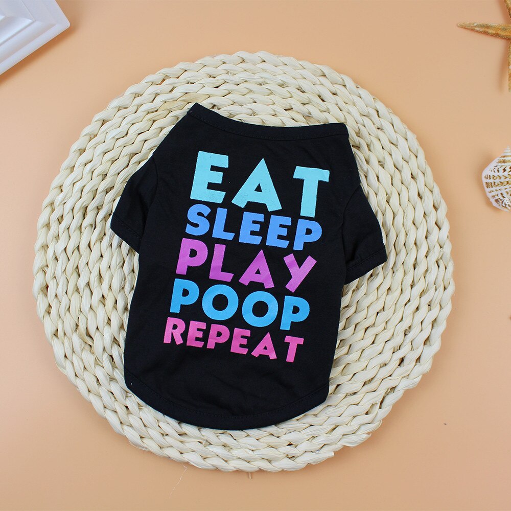 Black Eat Sleep Play Poop Repeat pet shirt laid flat on a woven mat with colorful lettering