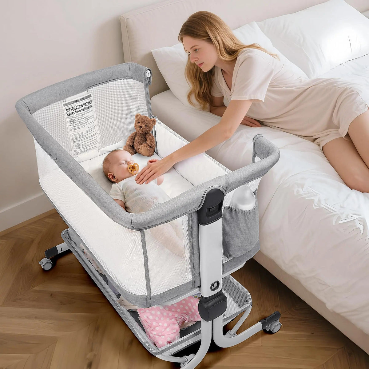 Woman leaning over a gray bedside bassinet with a baby and teddy bear inside.