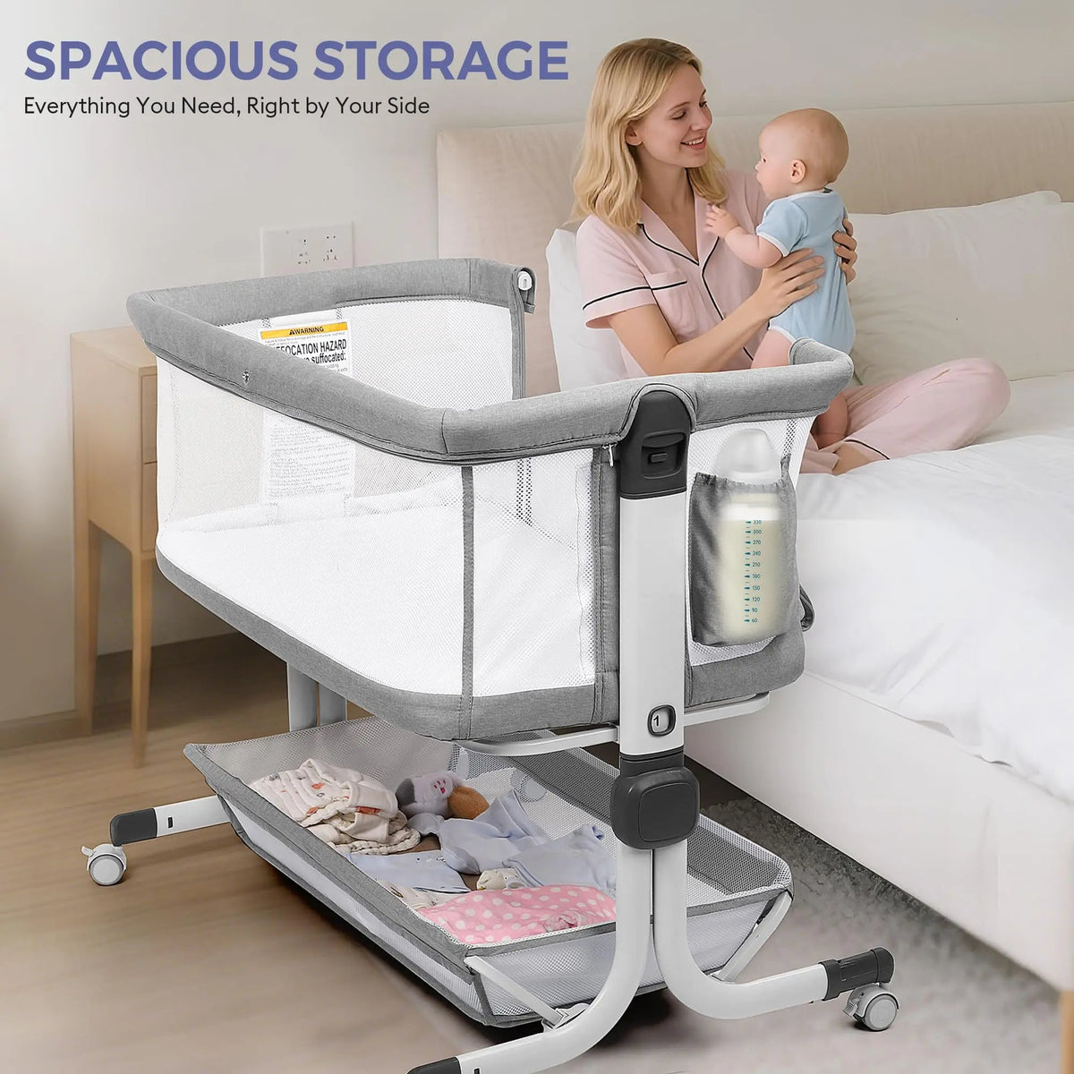 Mother holding a baby beside a gray bassinet with a side pocket and lower storage shelf.